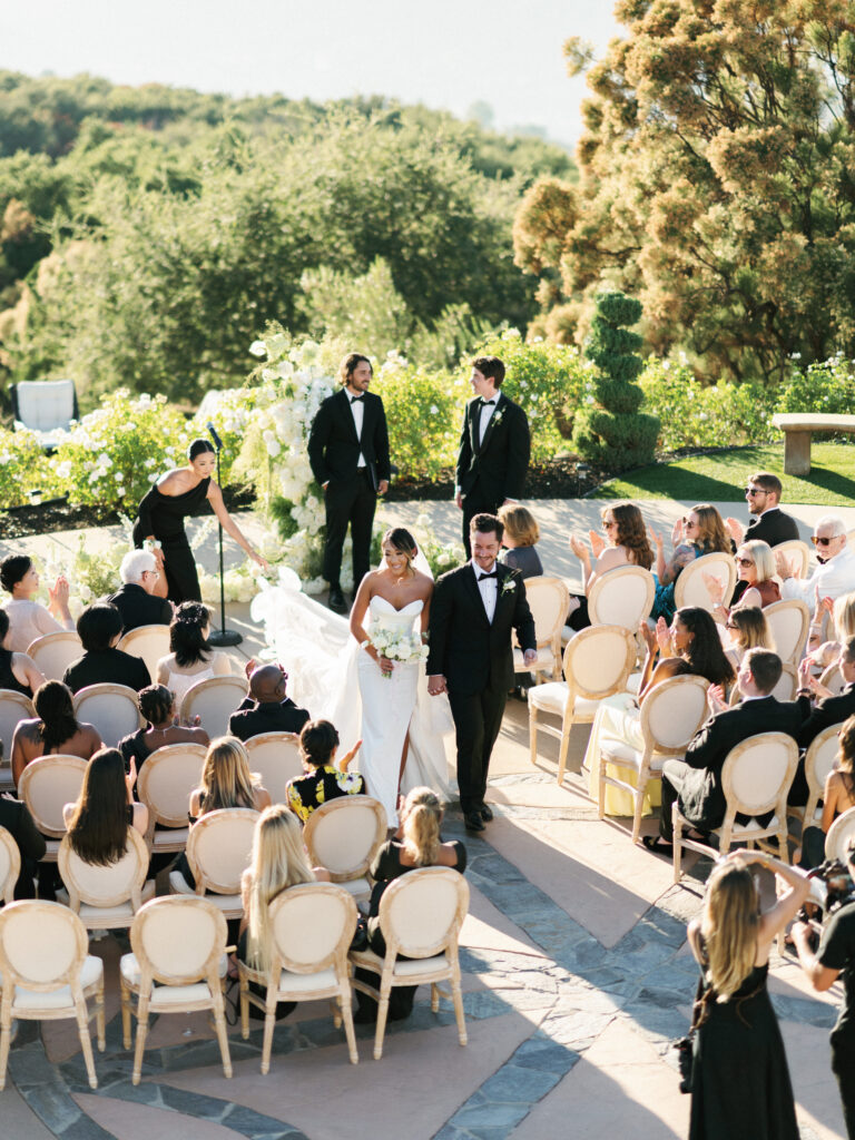 Bride and groom holding hands and walking down the aisle during a wedding ceremony recessional at the Formal Gardens of Greystone Mansion, with a coordinator adjusting the long dress train while guests applaud.