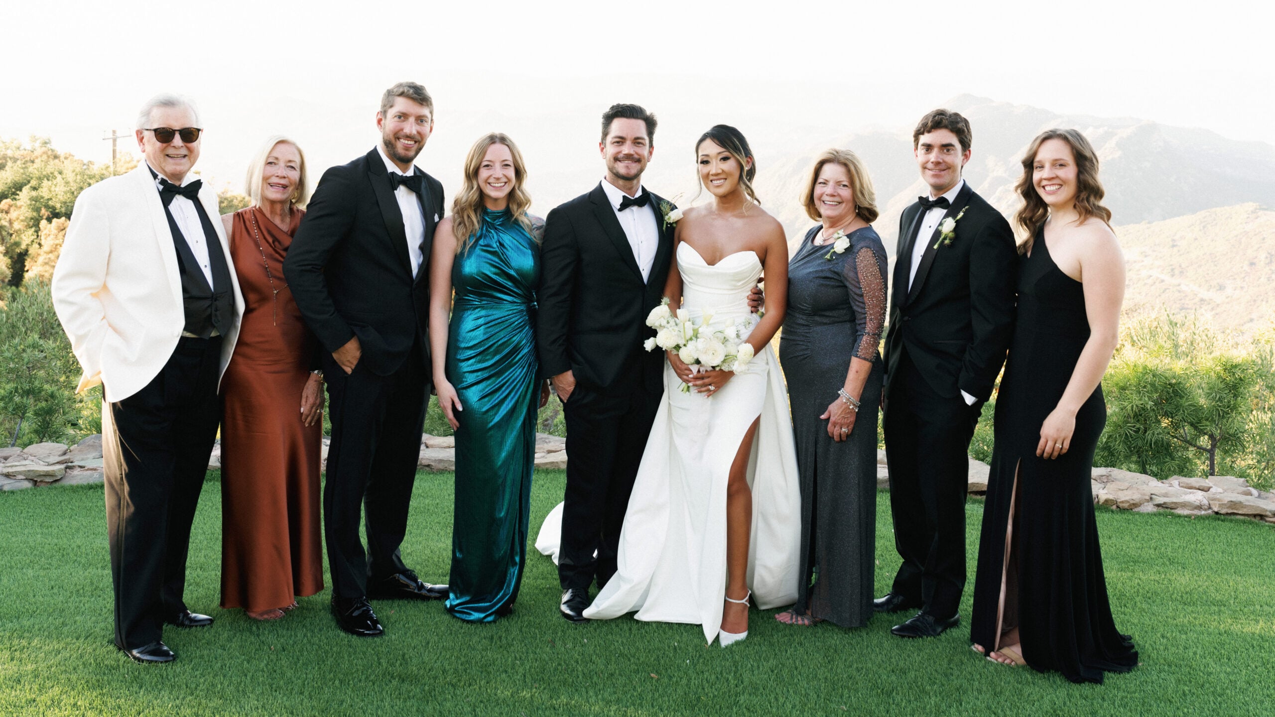 Editorial family wedding portrait on a scenic mountain overlook lawn in Malibu, featuring a modern bride in a high-slit gown and groom in a classic tuxedo with elegant guests.