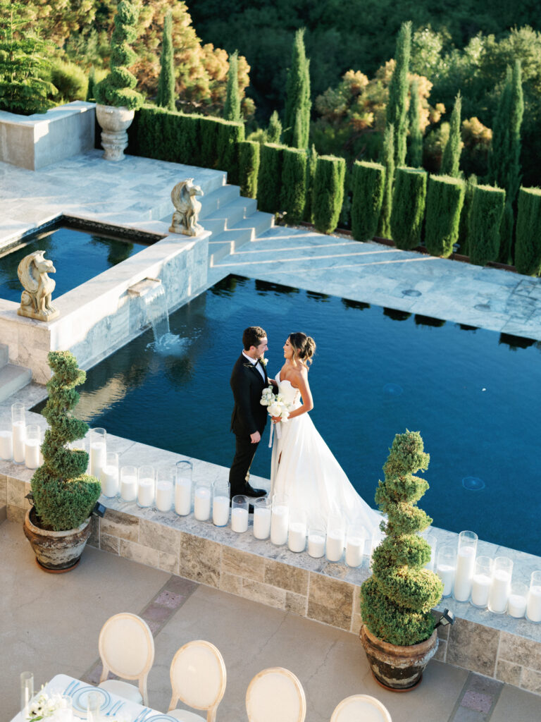 High-angle shot of a bride and groom standing together looking at each other by the reflection pool at the Pool Terrace of Greystone Mansion, surrounded by stone gargoyle statues, candles, and tall cypress trees.