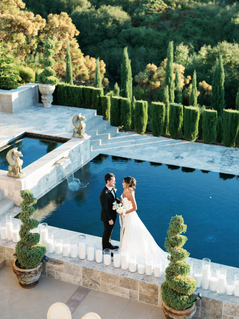 High-angle shot of a bride and groom looking at each other by the reflection pool at the Pool Terrace of Greystone Mansion, surrounded by stone gargoyle statues, cylinder candles, and tall cypress trees.