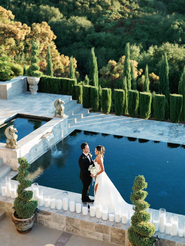 A high-angle view of a bride and groom standing by a luxury pool lined with glass cylinder candles at Cielo Farms in Malibu.