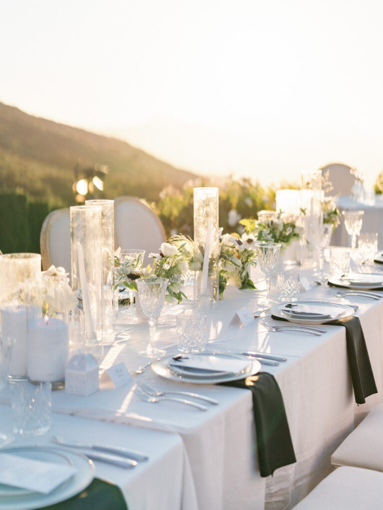 Close-up photography of an elegant wedding tablescape at the Pool Terrace of Greystone Mansion, featuring white tablecloths, glass cylinder candles, crystal glassware, and dark green napkins in golden hour light.