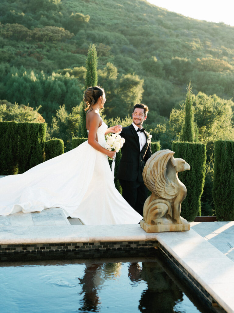 Bride in a strapless gown and groom in a tuxedo smiling at each other by the reflection pool at the Pool Terrace of Greystone Mansion, next to a historic stone griffin statue.