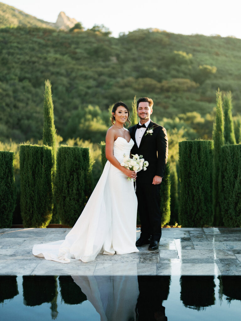 A bride and groom smile at a Cielo Farms wedding in Malibu, standing by a reflecting pool with cypress trees and mountains behind them.