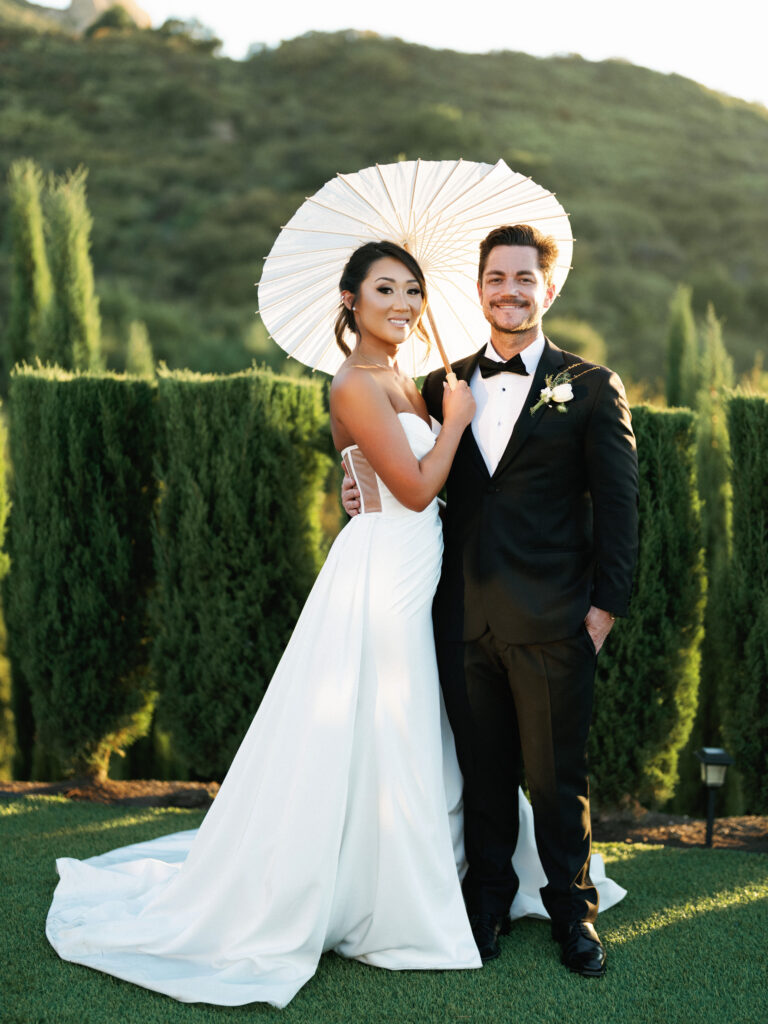 Bride holding a white paper parasol while standing with her groom on a green lawn at the Pool Terrace of Greystone Mansion, with tall hedges and sunny hills behind them.