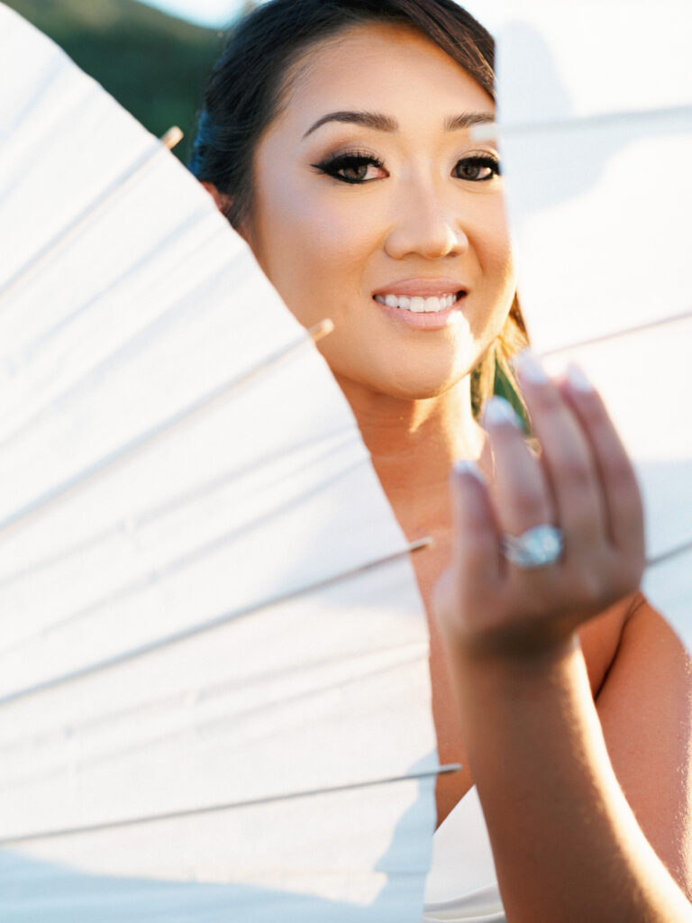 Macro portrait of a bride looking directly at the camera through a white sun umbrella, showcasing her elegant bridal styling and manicured hand with a diamond ring.