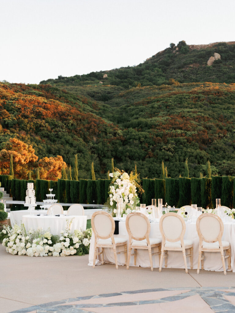 Wide shot of a luxury wedding head table layout at the Pool Terrace of Greystone Mansion, featuring round-back chairs, opulent white flower installations, and a multi-tiered wedding cake against a forested hill.