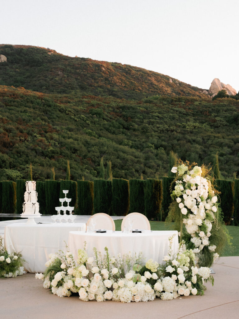 Elegant outdoor wedding reception table heavily decorated with white roses and hydrangeas, next to a tiered wedding cake and a pyramid of champagne glasses under a clear sky.