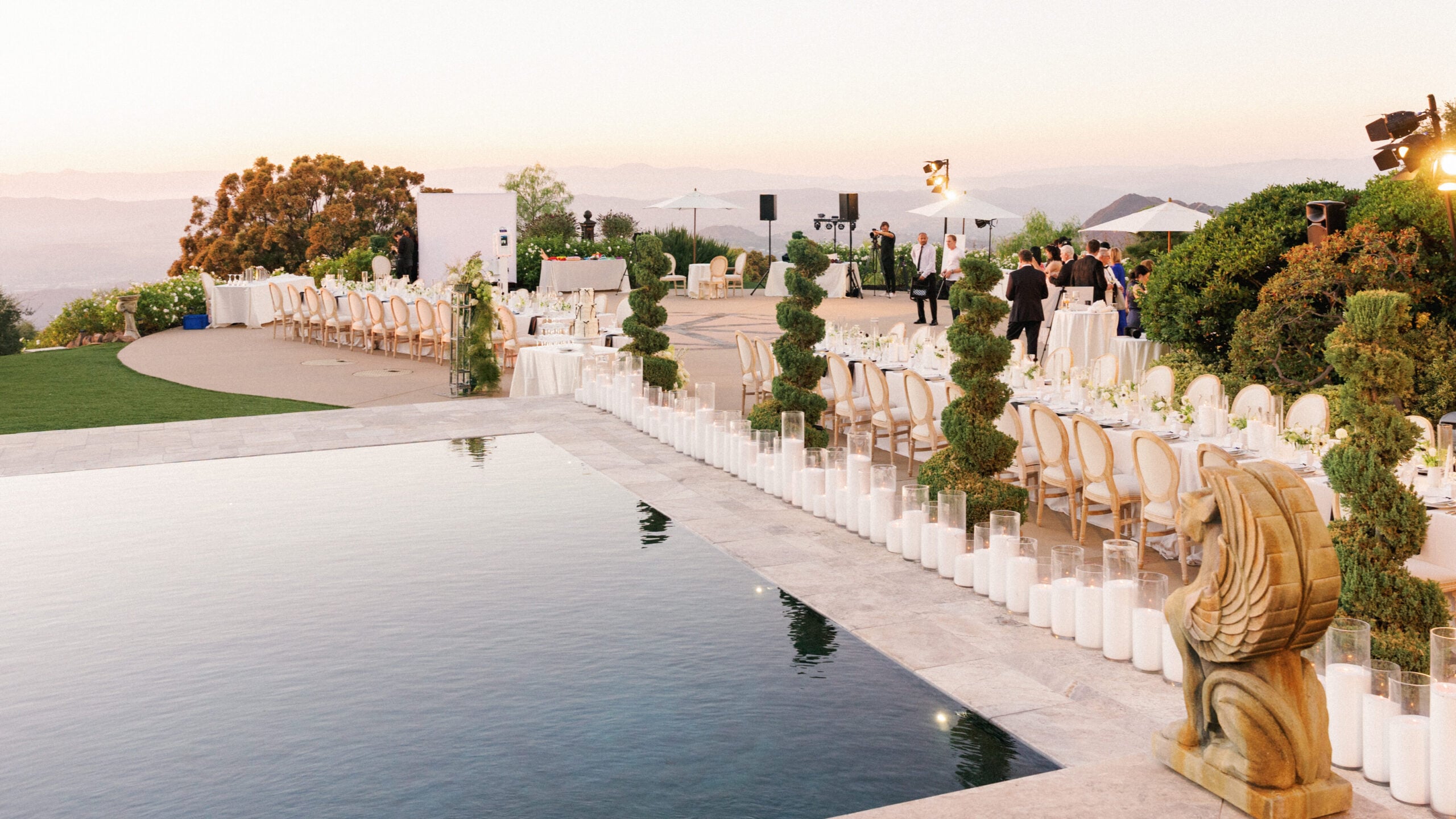 Wide shot of an outdoor luxury wedding reception setup at the Pool Terrace of Greystone Mansion, featuring a long row of glass cylinder candles by a reflection pool, banquet tables with round-back chairs, spiral topiary trees, and a stone griffin statue during sunset.
