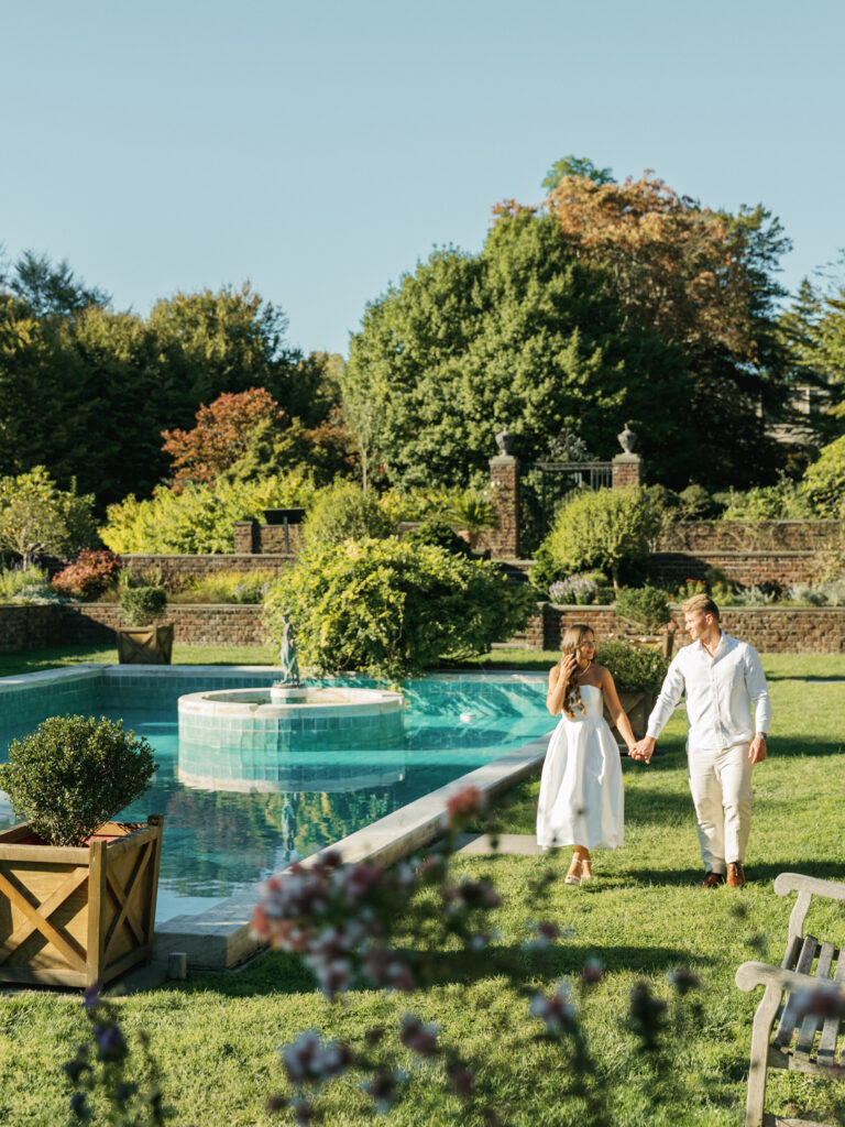 Future bride in a strapless white midi dress and groom in a light linen shirt walking hand in hand by a turquoise fountain pool at a historic estate garden.