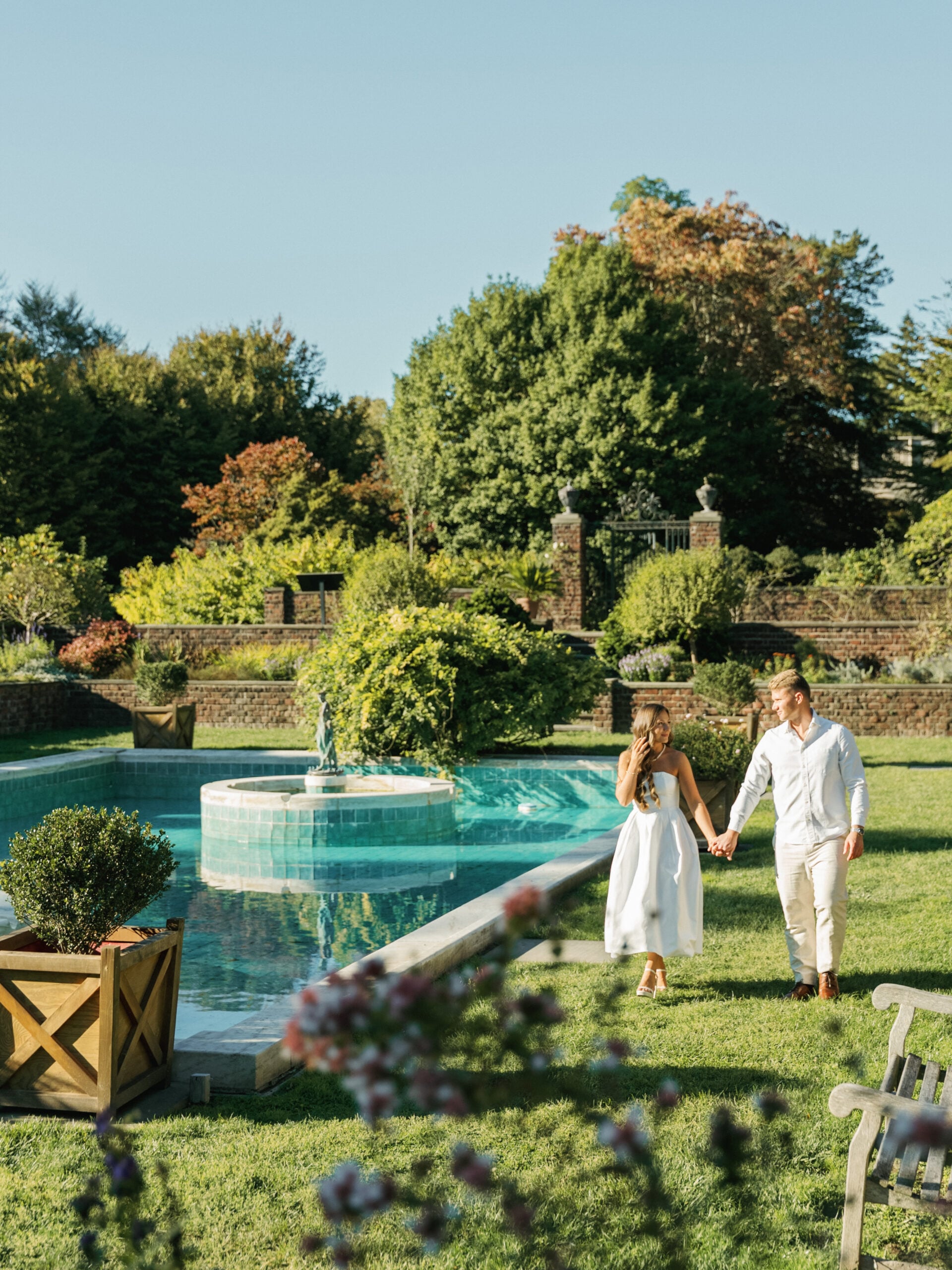 Future bride in a strapless white midi dress and groom in a light linen shirt walking hand in hand by a turquoise fountain pool at a historic estate garden.