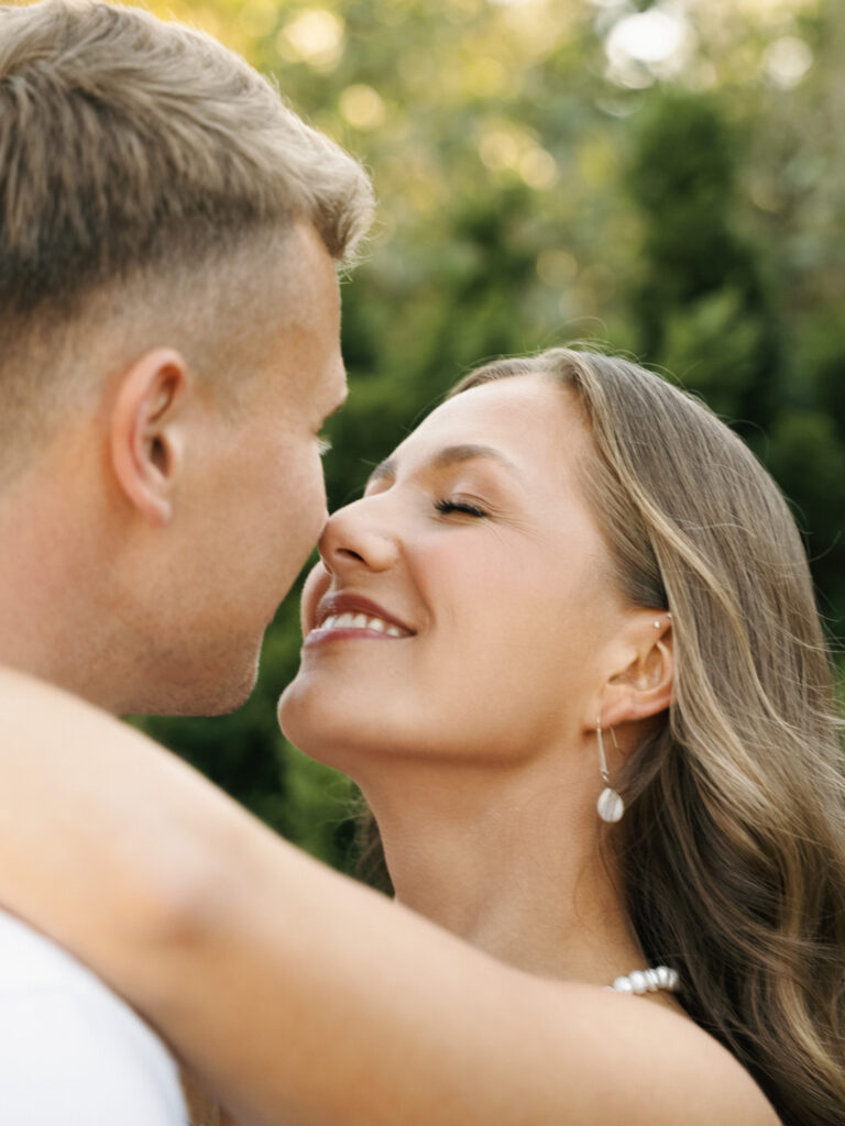 Close-up portrait of a smiling bride wearing a pearl necklace and drop earrings hugging her groom outdoors during a sunset shoot.