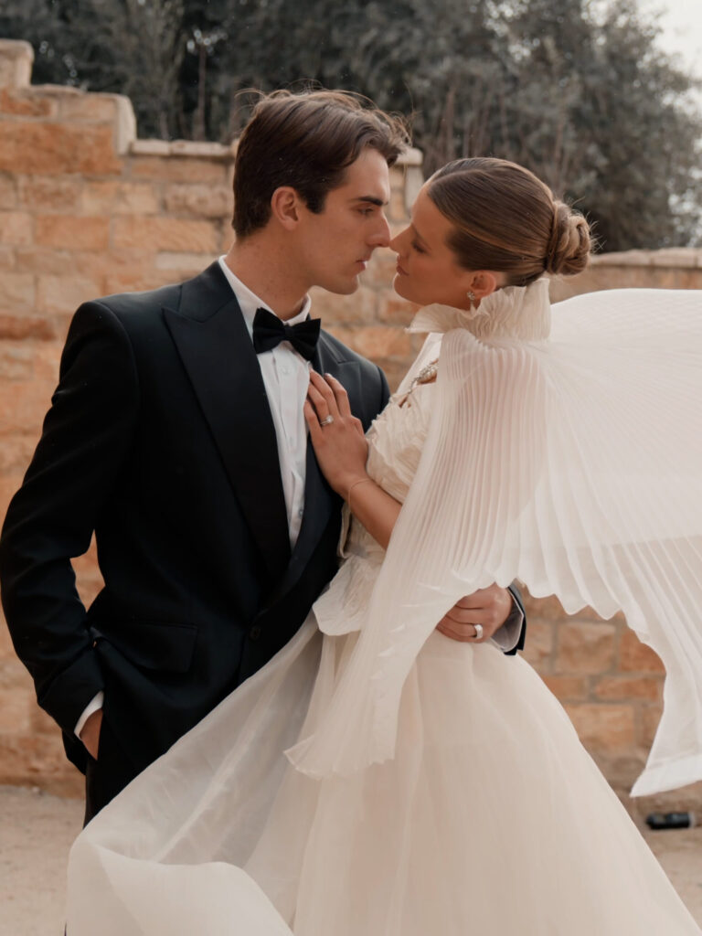 Medium close-up profile of a bride and groom embracing by a rustic stone wall at Sunstone Winery, featuring the bride in a high-fashion pleated cape dress with her hand on the groom's chest.