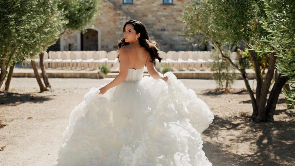 Editorial back view of a bride running on a gravel courtyard toward an outdoor ceremony setup at Sunstone Villa, lifting her dramatic textured ruffle ballgown and looking over her shoulder.