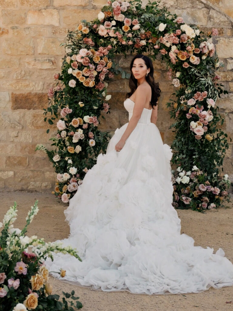 High-fashion bridal portrait at Sunstone Winery, showing the bride in a dramatic tulle ruffle wedding dress with a long train posing in front of a lush, romantic blush and mauve floral arch.