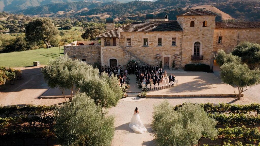 Bride walking down the aisle during an outdoor wedding ceremony processional at Sunstone Villa, featuring a long olive tree pathway and mountain views in Santa Ynez.