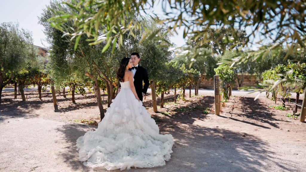 Elegant couple portrait at Sunstone Villa wedding, featuring a bride in a luxury textured ruffle ballgown and a groom in a classic black tuxedo among Santa Ynez olive trees and vineyards.