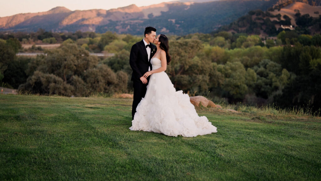 Bride and groom kissing during sunset wedding photos at Sunstone Villa, showcasing a luxury ruffle ballgown on a green lawn with Santa Ynez mountain views.