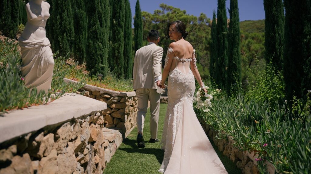 Bride and groom holding hands and walking away along the Cypress Walk at Greystone Mansion, with the bride looking back over her shoulder during a photo and film portrait session.