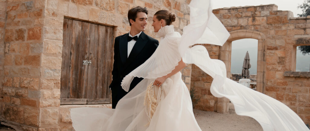 Three-quarter view of a bride and groom on a stone courtyard at Sunstone Villa, showcasing a modern high-neck wedding dress with a long pleated cape blowing in the wind next to historic stone walls.