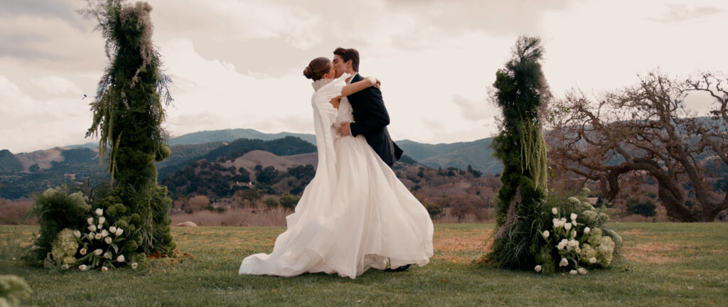 Editorial wedding photography of a bride in a couture cape dress and groom kissing at their Sunstone Villa ceremony, featuring organic moss and white tulip floral pillars with Santa Ynez mountains view.