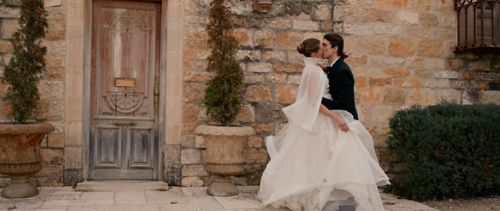 Bride and groom kissing against the rustic stone walls of Sunstone Villa, showcasing a modern wedding dress with a pleated cape next to a historic carved wooden door.