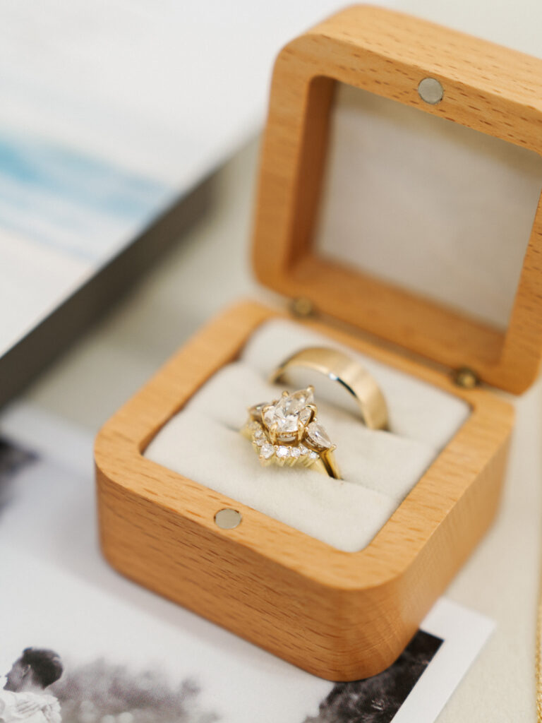 Unique pear cut diamond engagement ring and thick gold wedding band resting in a light wood square ring box with a white velvet cushion over a black and white wedding photo.