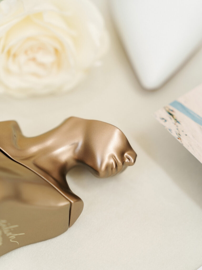 Billie Eilish metallic bronze bust perfume bottle lying flat on a beige linen surface next to a white garden rose petal and blurred bridal heels.