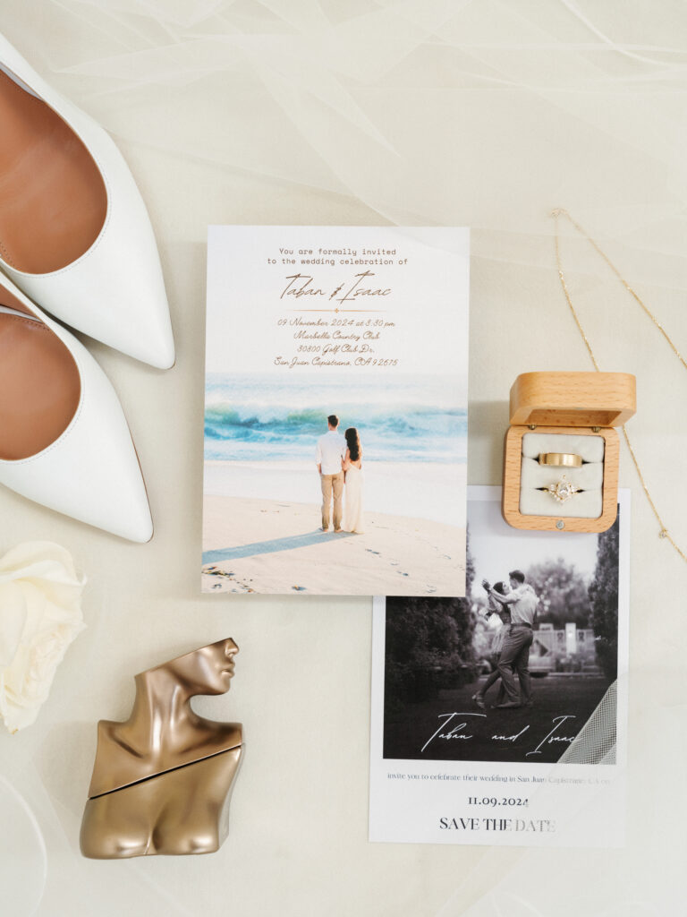 Beach destination wedding invitation suite flat lay with a coastal photo save the date card, white leather pointed toe flats, Eilish perfume, and a wooden ring box on a white tulle backdrop.
