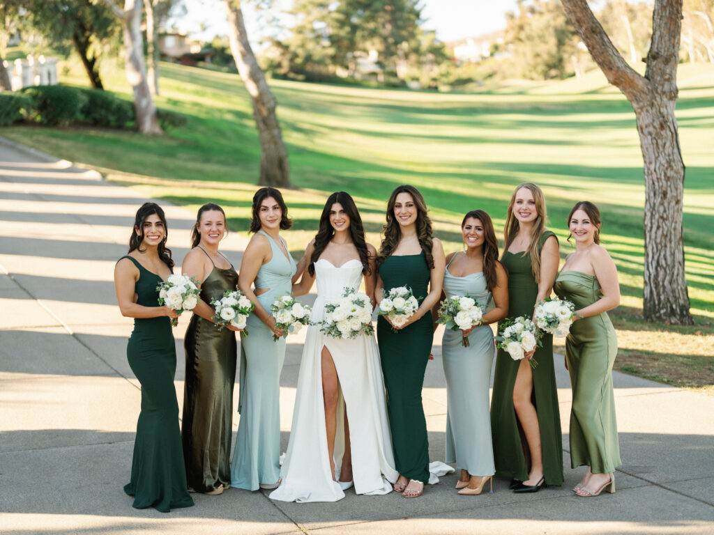 Bride in a strapless high-slit gown posing with bridesmaids in mismatched green, sage, and emerald silk dresses holding white rose bouquets at an outdoor country club wedding.