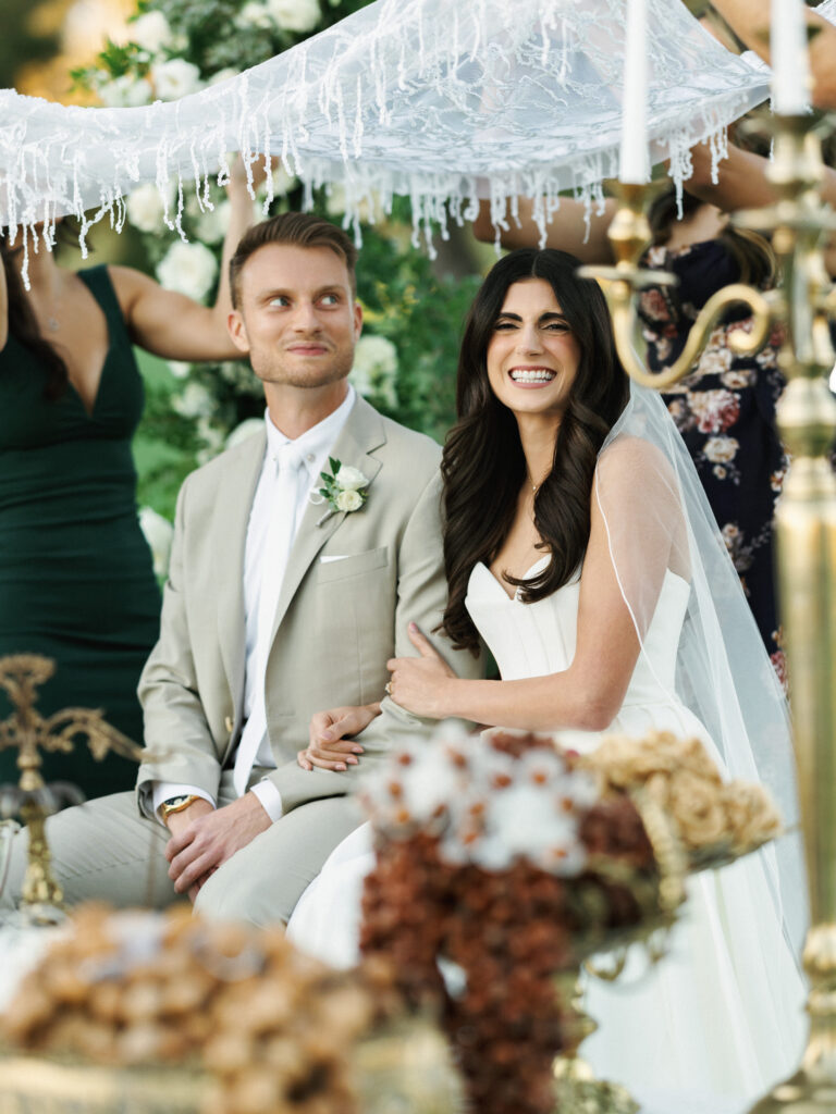 A bride and groom in a classic white dress and beige suit sit together during a Persian Sofreh Aghd ceremony as part of their multicultural and traditional celebration.