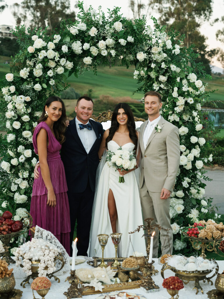 Sophisticated bridal party portrait featuring the newlyweds with their elegant wedding guests on a scenic garden lawn under a magnificent floral installation.