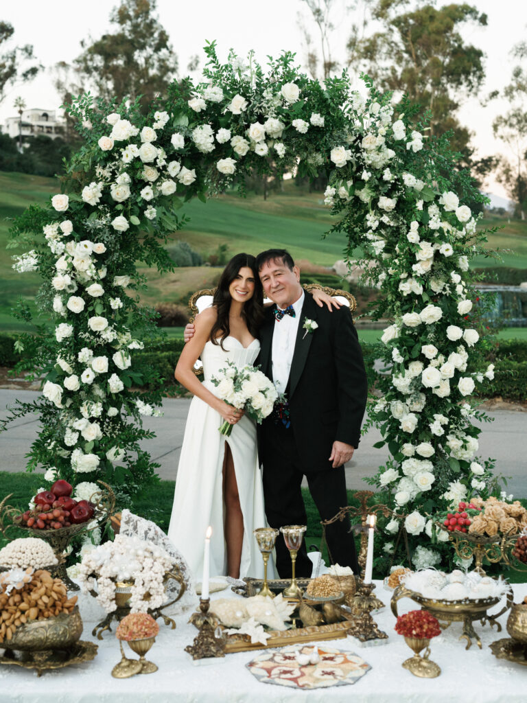 Elegant bride in a modern high-slit wedding gown posing with her father under a lush white rose floral arch at an outdoor California estate ceremony.