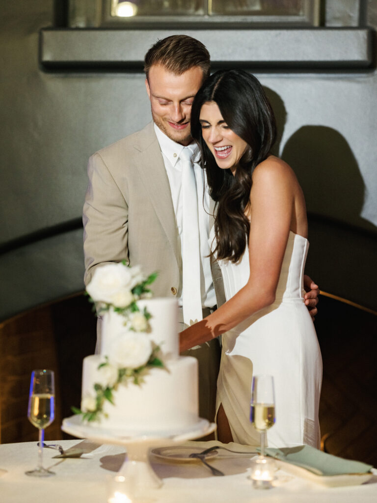 Happy newlyweds laughing while cutting an elegant tiered wedding cake decorated with fresh white flowers.