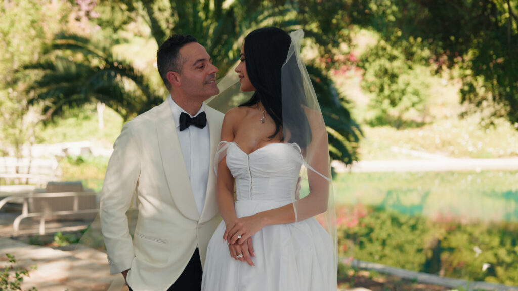 Cinematic wedding videography style newlyweds portrait, bride in a corset couture gown and groom in a white tuxedo look at each other, luxury estate garden.