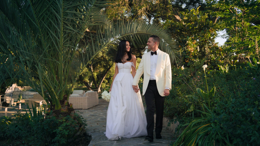 California outdoor black tie wedding ideas, bride in a corset gown and groom in a white tuxedo jacket walking by palm trees.