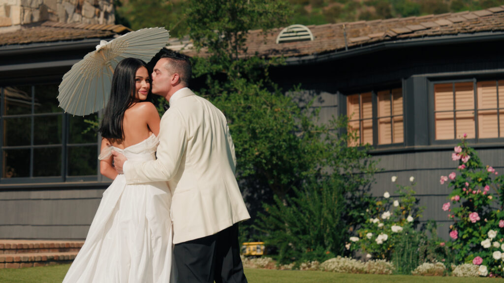 Luxury backyard estate wedding, bride in a silk off-the-shoulder wedding dress holding a paper umbrella with groom in a white dinner jacket.