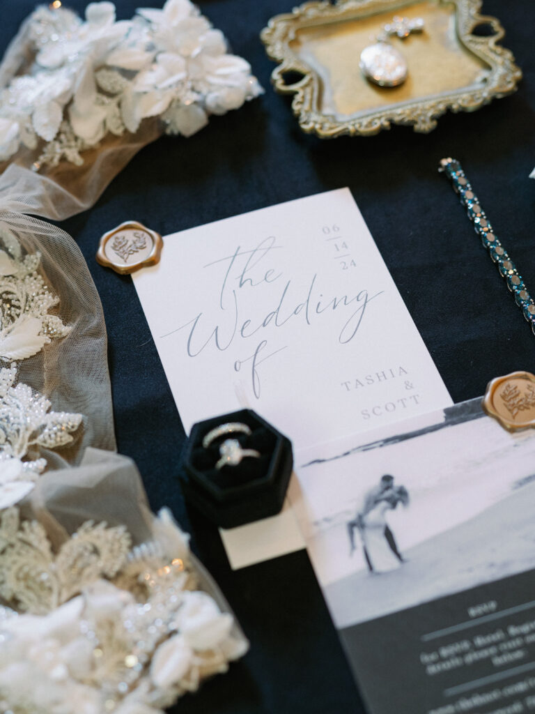 Modern black and white wedding invitation flat lay with minimalist calligraphy text, wax seals, a black velvet ring box, and embroidered lace details.