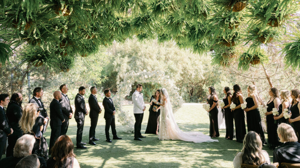 An outdoor California wedding ceremony with a lush hanging fern canopy, a white circular floral arch, and a bridal party in black tie.