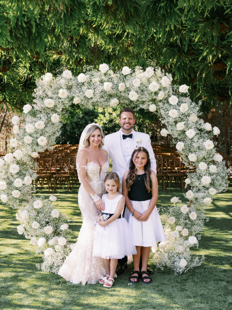 Bride in a lace gown and groom in a white tuxedo posing with two flower girls in white and black dresses in front of a circular baby's breath wedding arch.