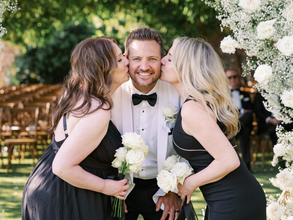 Groomsmen or family members giving a candid cheek kiss to a groom in a white tuxedo jacket holding white roses at an outdoor wedding.