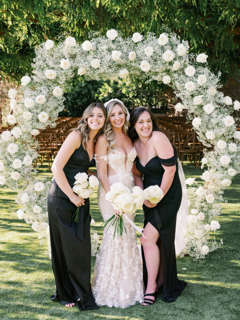 Modern outdoor wedding ceremony portrait featuring a bride in a long veil with two bridesmaids in formal black dresses next to a large round floral installation.