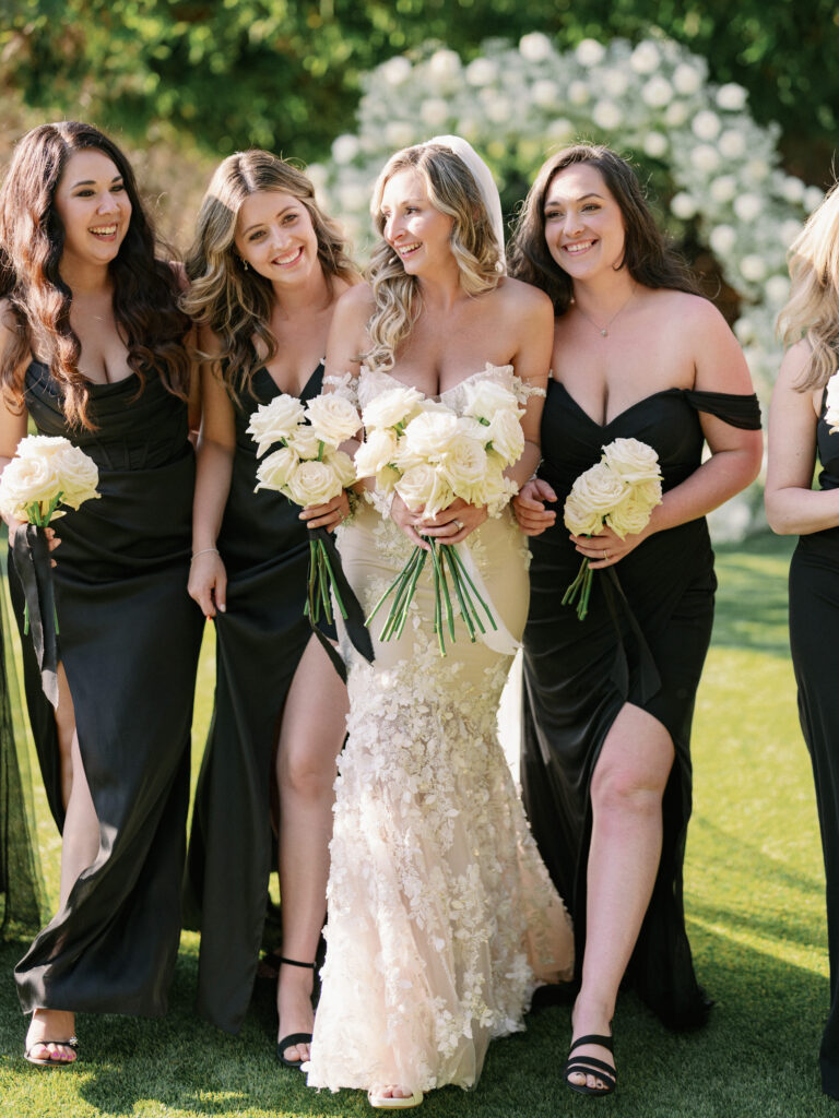 Smiling bride in an off-the-shoulder lace mermaid gown walking arms linked with bridesmaids in black satin slit dresses holding white rose bouquets.