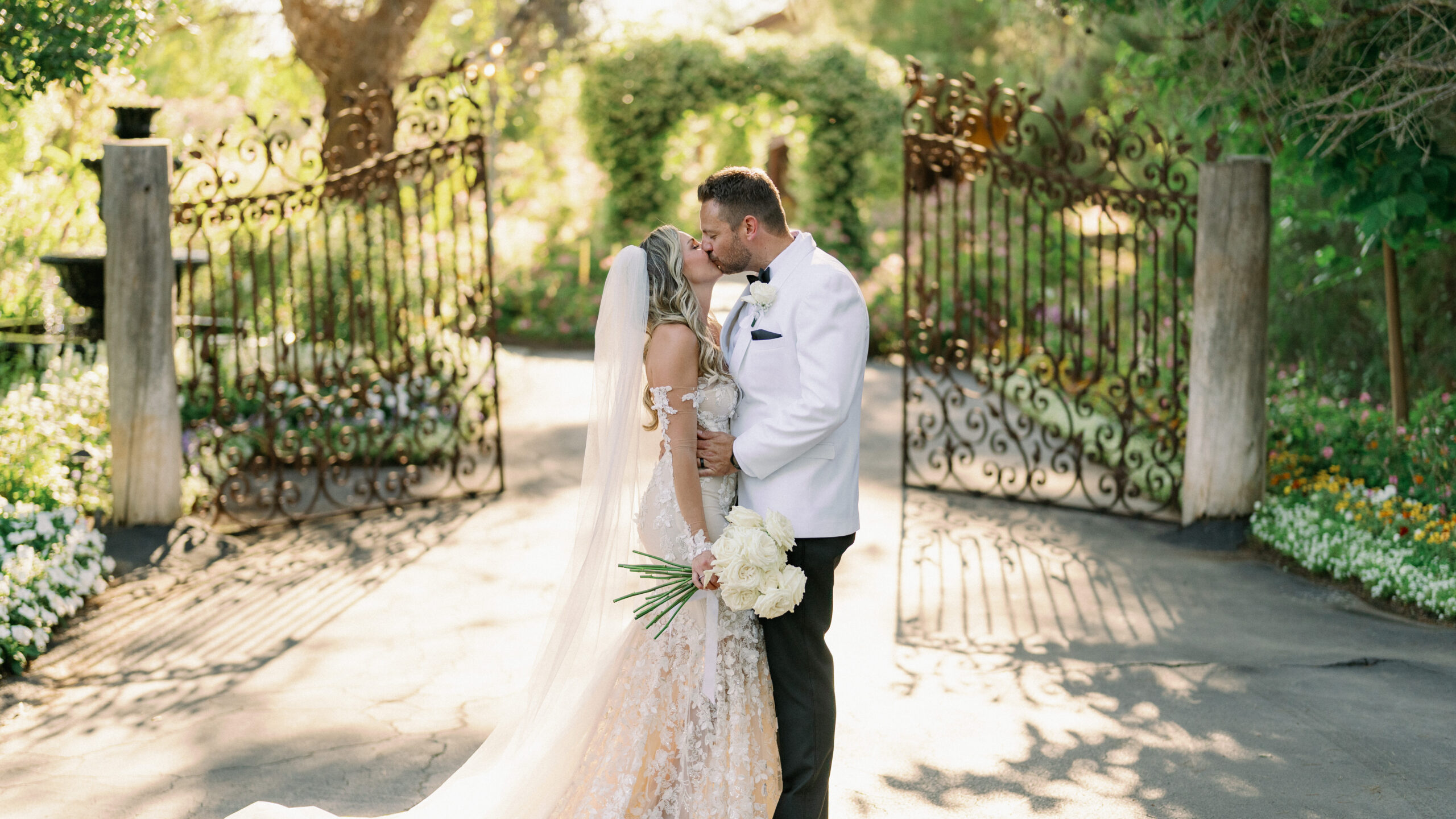 Elegant California wedding photography of a groom in a white tuxedo jacket and a bride with a long cathedral veil holding a white rose bouquet.