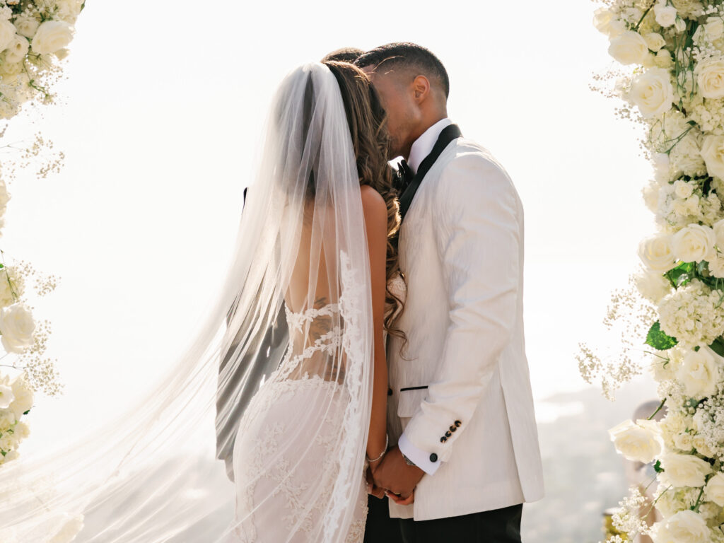 Bride in a backless lace couture wedding gown and long tulle veil kissing groom in a white tuxedo jacket, luxury cliffside estate ceremony destination wedding.