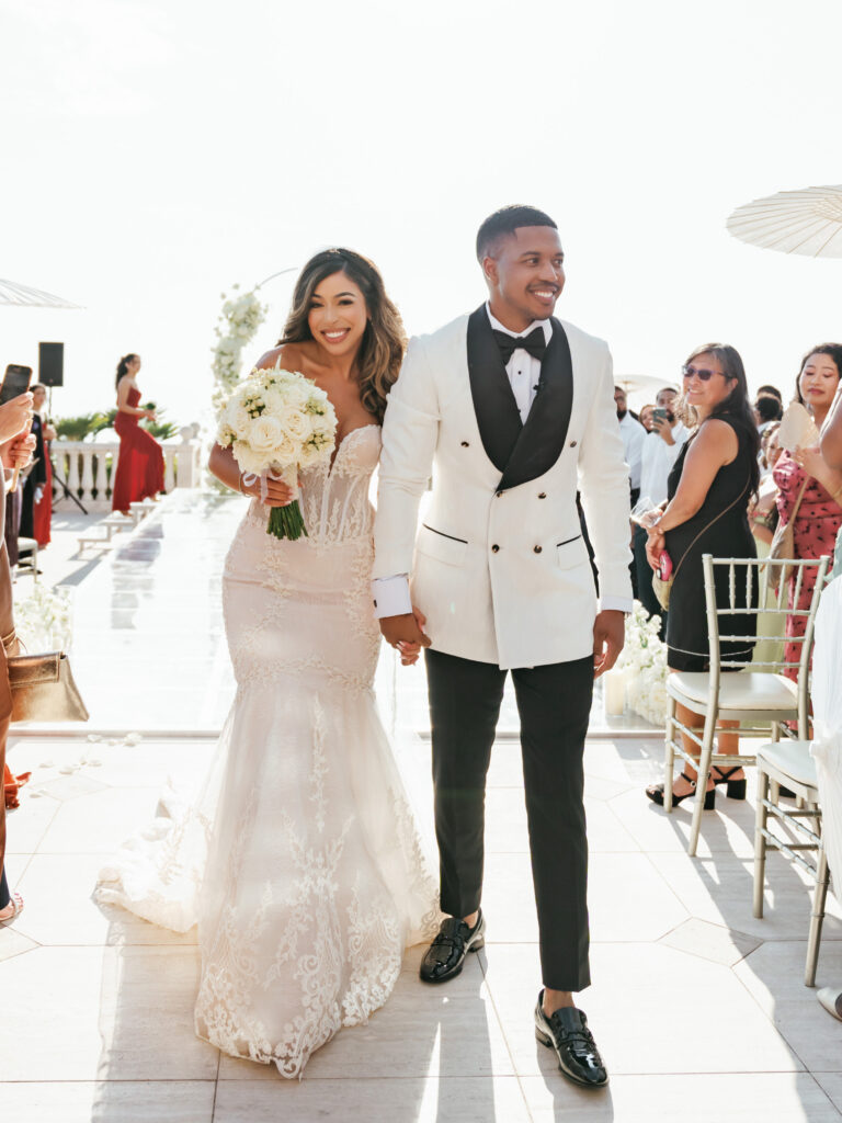 Bride in an embellished mermaid lace gown and groom in a bespoke white double-breasted tuxedo walking together, high-fashion outdoor wedding inspiration.