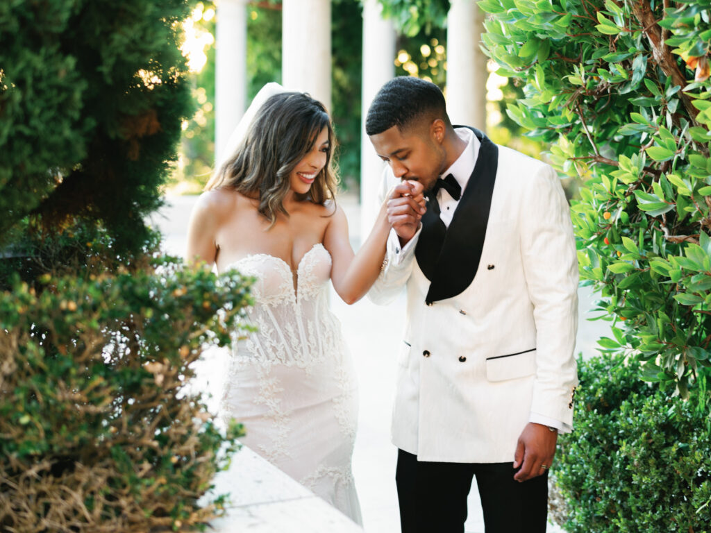 Groom in a white bespoke tuxedo kissing hand of a bride in a strapless lace corset gown, sophisticated editorial wedding photography.