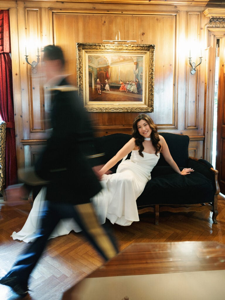 Motion blur wedding photography of a bride in a modern gown sitting on a vintage sofa with a groom walking past in a wood-walled estate room.