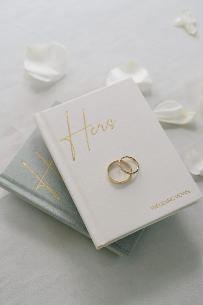 Elegant white and grey velvet wedding vow books with gold foil calligraphy, holding two gold wedding bands with white rose petals around.