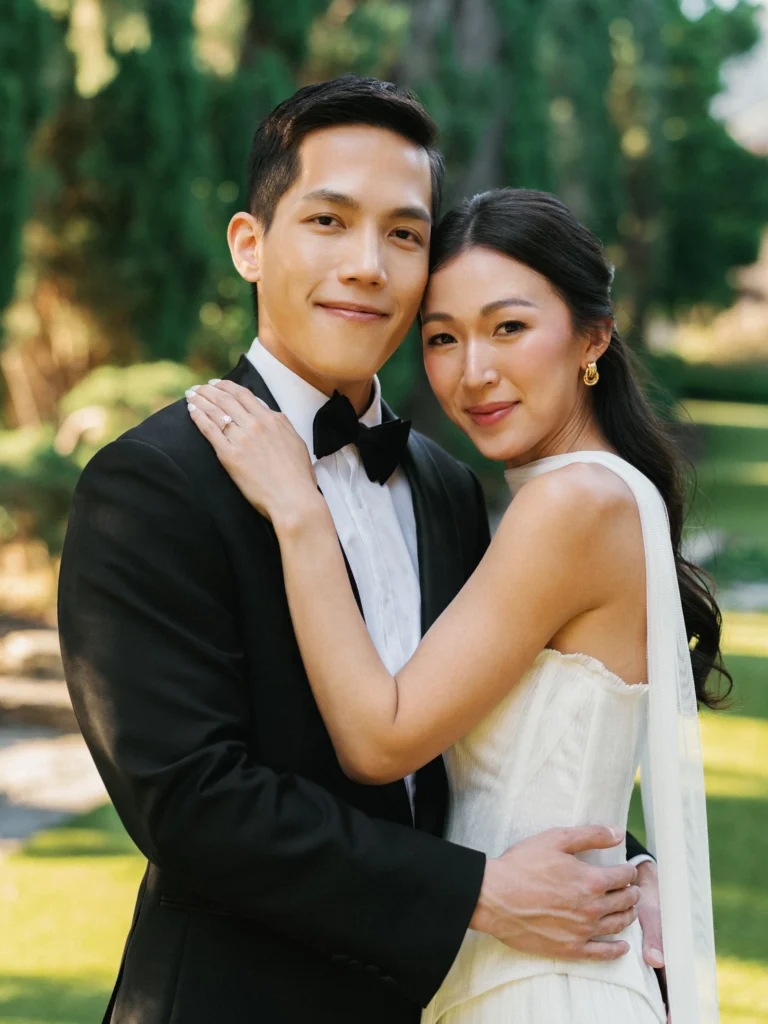 Vertical medium shot of a wedding couple embraced on a green lawn, highlighting the bride's half-up wavy hairstyle and the groom's classic black suit.